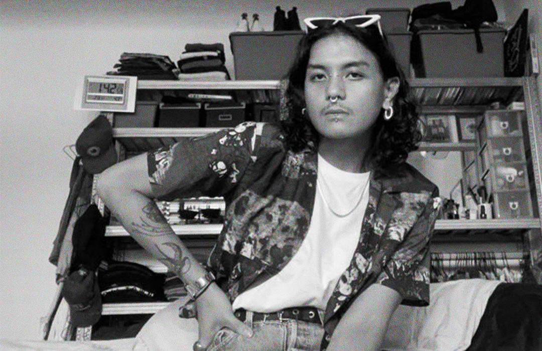 Bryan is sitting down facing the camera, wearing a patterned shirt and sunglasses sitting in front of a shelf with folded clothes.