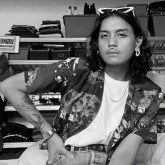 Bryan is sitting down facing the camera, wearing a patterned shirt and sunglasses sitting in front of a shelf with folded clothes.